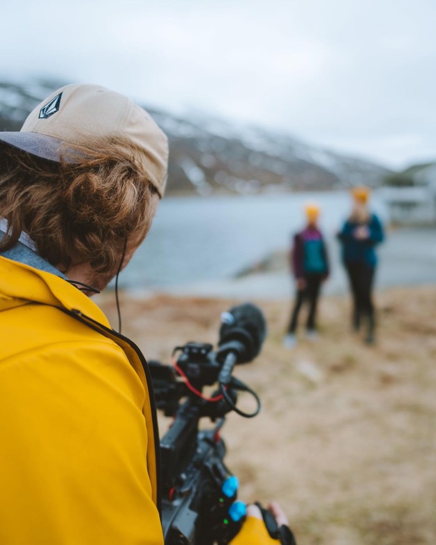 A photographer dressed in a yellow jacket, with a caps backwards. He is filming two girls who are far away and blurry.